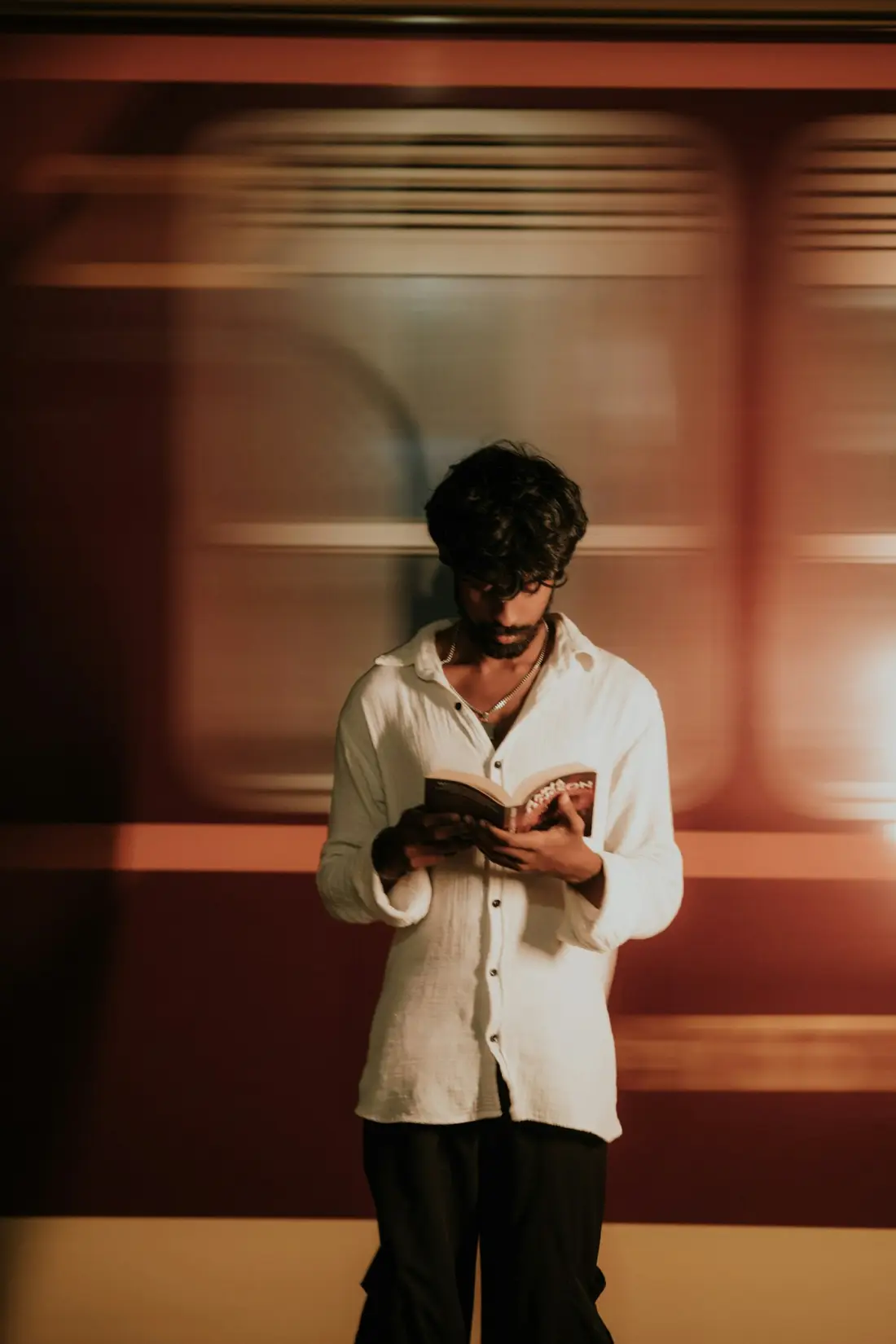 A man in a white shirt reads a paperback on a platform as a train blurs past behind him